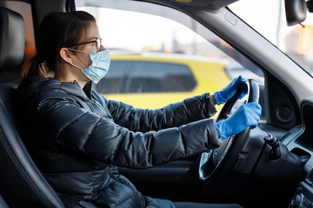 Mujer bastante caucásica con gafas, máscara médica antivirus y guantes de látex azul conduciendo un automóvil