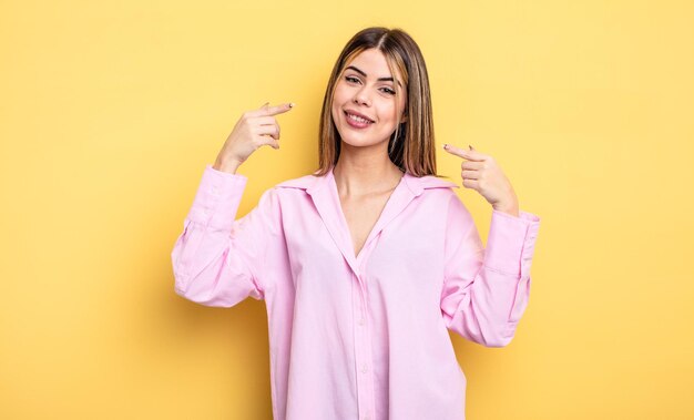 Mujer bastante caucásica sonriendo con confianza apuntando a su propia sonrisa amplia, actitud positiva, relajada y satisfecha