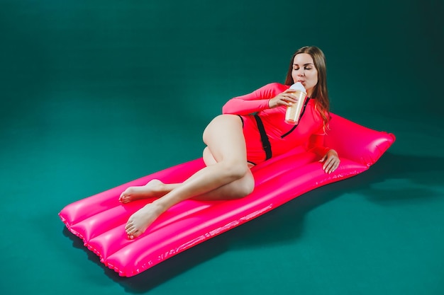 Mujer bastante delgada en traje de baño rosa yace sobre un colchón inflable con un cóctel en las manos sobre un fondo verde aislado Vacaciones de verano en el concepto de bronceado en el mar Colchón de aire rosa