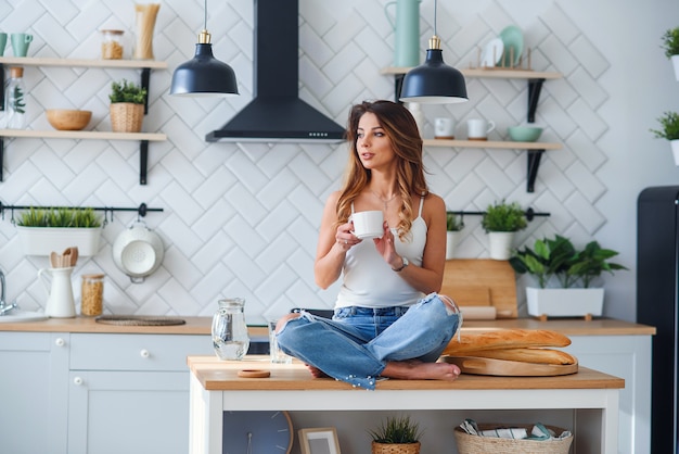 La mujer bastante feliz bebe café mientras está sentado en la mesa en la acogedora cocina por la mañana en casa. Relajación matutina en casa.