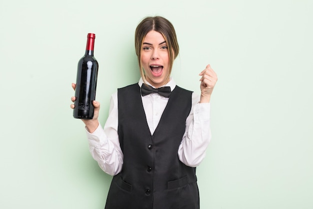 Mujer bastante joven que se siente sorprendida, riendo y celebrando el concepto de camarero y botella de vino de éxito