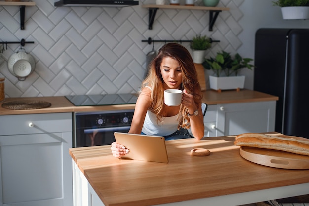 Mujer bastante joven que usa la tableta mientras que bebe el café después de desayuno en la cocina por la mañana.