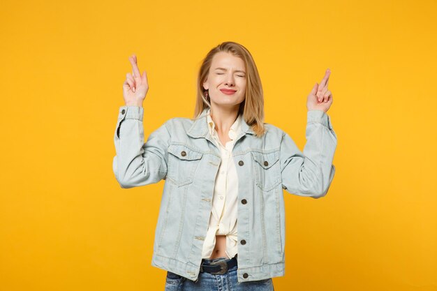 Mujer bastante joven con ropa de mezclilla esperando un momento especial, con los dedos cruzados, los ojos cerrados, pidiendo deseos aislados en un fondo amarillo anaranjado. Concepto de estilo de vida de las personas. Simulacros de espacio de copia.