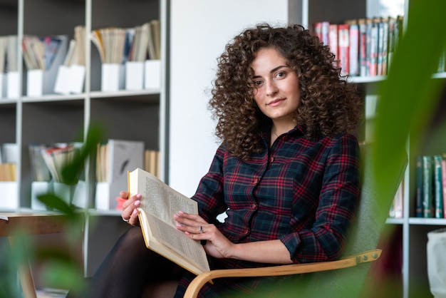 La mujer bastante joven se sienta en la silla y sostiene el libro, mirando la cámara. Libro abierto sobre la rodilla- Imagen