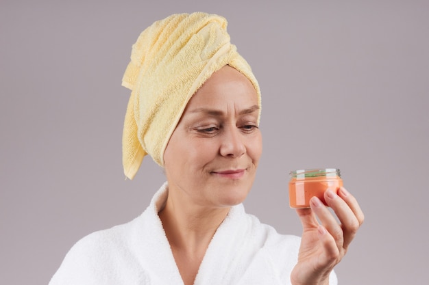 Una mujer bastante madura con una bata, una toalla amarilla en la cabeza, sosteniendo un tarro de crema de naranja. Concepto de cuidado de la piel facial. sobre la pared gris.