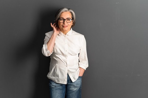 La mujer bastante madura corrige el peinado de longitud media coquetamente. Adorable modelo caucásico de pelo gris en jeans posando sobre fondo gris. Copiar espacio o espacio de texto en el lado derecho