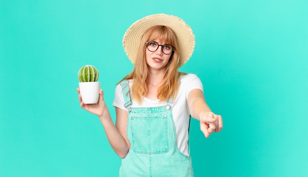 Mujer bastante pelirroja apuntando a la cámara eligiéndote y sosteniendo un cactus en maceta. concepto de granjero