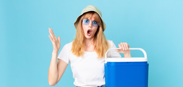 Mujer bastante pelirroja que se siente extremadamente conmocionada y sorprendida y sosteniendo un refrigerador portátil. concepto de verano