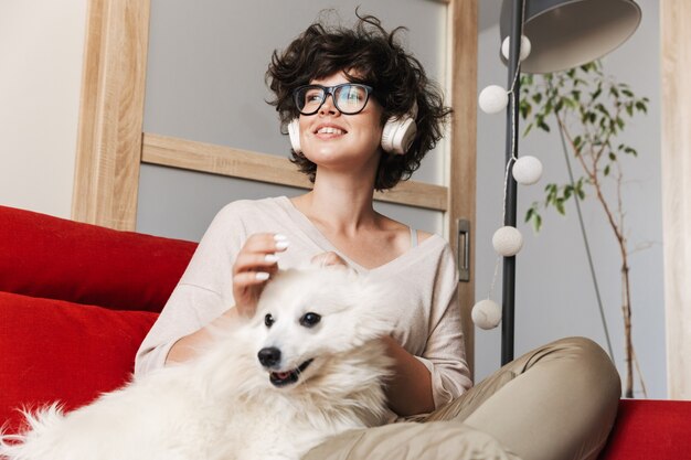 una mujer bastante rizada sentada en el sofá con su lindo perro blanco escuchando música con auriculares.