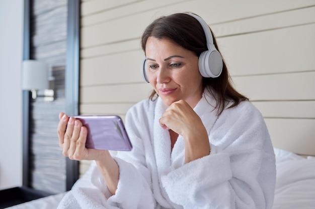 Mujer en bata de baño y auriculares mirando la pantalla del teléfono inteligente sentada en la cama