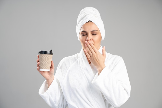 Mujer en bata de baño blanca con toalla en la cabeza sosteniendo una taza de té o café en la mano y bostezando después de la ducha por la mañana. Rutina diaria de la mañana y comienzo de un nuevo concepto de día.