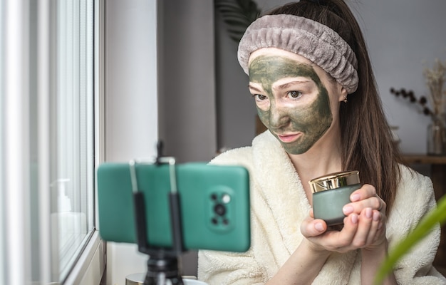 Mujer en bata de baño con una máscara cosmética verde en su rostro está mostrando un frasco de máscara en un teléfono móvil