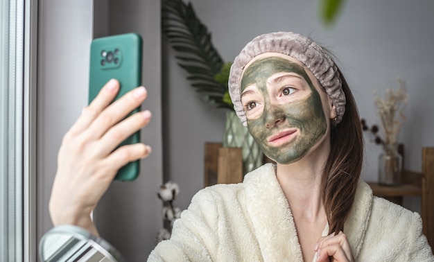 Mujer en bata de baño y con una máscara cosmética verde en su rostro está tomando un selfie en su teléfono