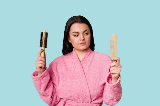 Una mujer con una bata de baño rosa sosteniendo un cepillo y un peine con una toalla envuelta alrededor de su cabeza