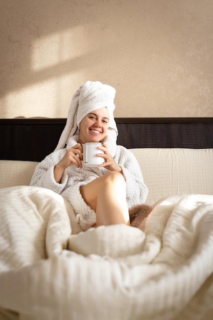 Una mujer con una bata blanca y una toalla en la cabeza se sienta en una cama y sostiene una taza