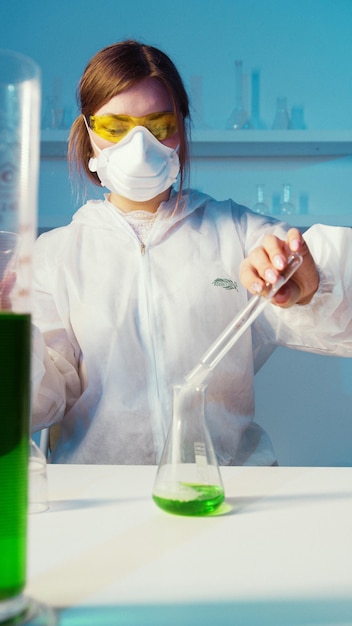 Una mujer con bata de laboratorio y gafas amarillas vierte líquido en un vaso de precipitados.