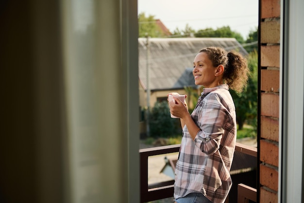 Mujer bebe café admira el paisaje rural desde el balcón de la casa de campo en un día soleado