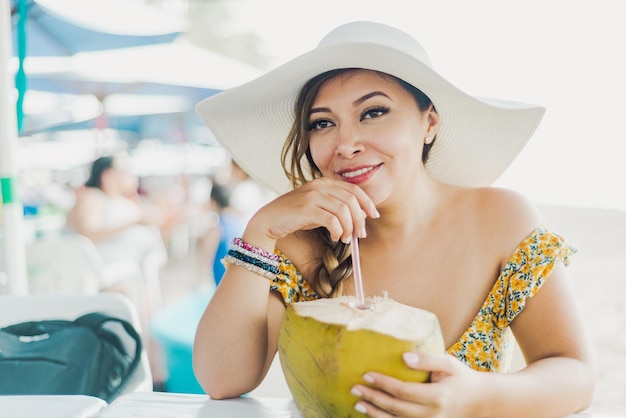 Mujer bebiendo agua de coco. Mujer con sombrero de playa bebiendo agua de coco con una pajita.