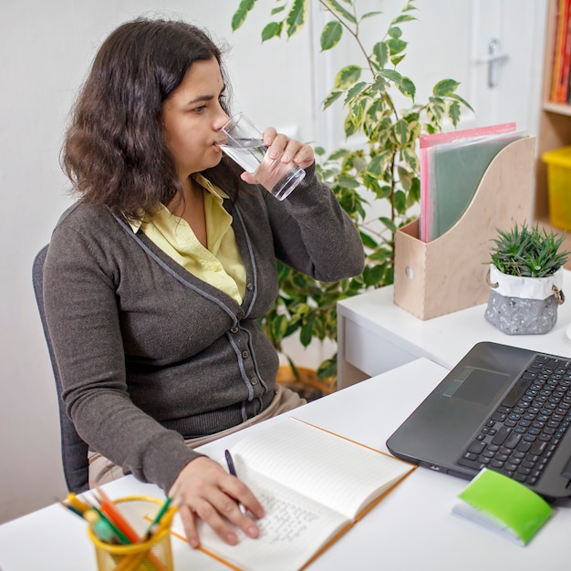 Mujer bebiendo agua en la oficina delante de la computadora portátil