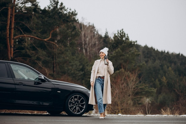 Mujer bebiendo café junto a su coche de pie en la carretera en el bosque