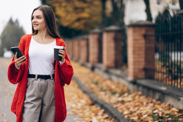 Mujer bebiendo café y usando el teléfono y caminando en la calle de otoño