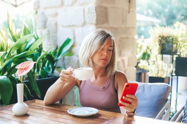 Mujer bebiendo su té mientras usa un teléfono inteligente para mensajes sms, escribiendo un teléfono celular con pantalla táctil en el café soleado. Estilo de vida, tecnología y concepto de red de medios sociales.