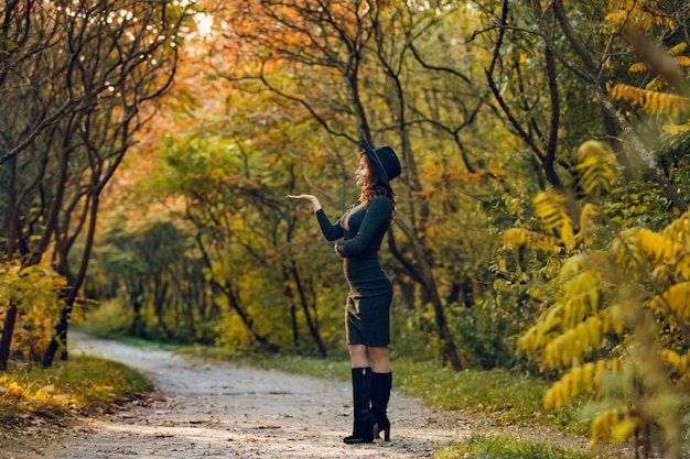 Una mujer con una bella figura en un sombrero negro se para de lado en el parque y posa para una sesión de fotos