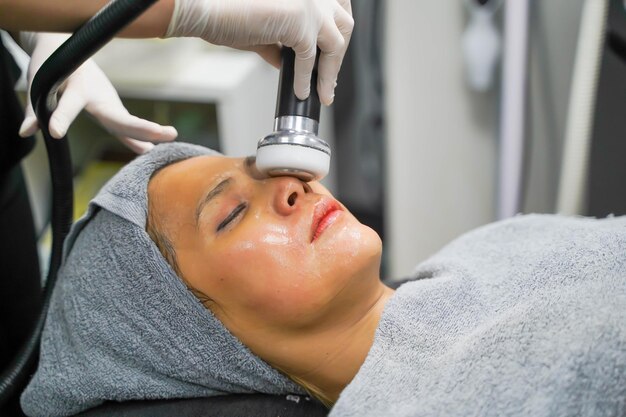 Mujer de belleza asiática de primer plano que recibe terapia para estimular la piel facial y el tratamiento ultrasónico facial para el cuidado de la piel por parte de una cosmetóloga profesional