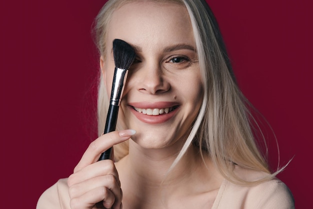 Mujer de belleza con cubierta de cepillo en la cara para polvo cosmético o herramientas de base Cosméticos de maquillaje y cepillos para modelo de niña hermosa aislado sobre fondo de color cereza