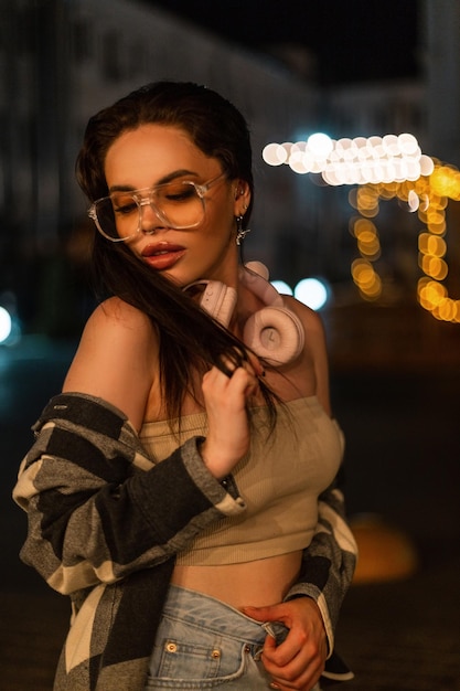 Mujer de belleza con gafas de moda y auriculares en un elegante atuendo casual camina por la ciudad por la noche