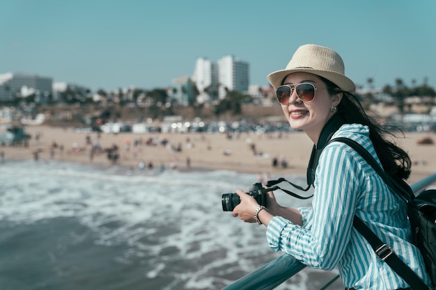 Mujer de belleza hace una foto de la playa del océano mientras está de pie en el muelle apoyándose en la barandilla. Fotografía y concepto de estilo de vida de viajes. Joven dama elegante con sombrero de paja sosteniendo una cámara sonriendo disfrutando de la vista de la naturaleza