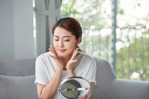 Mujer de belleza mira mirrior felizmente y toca su rostro