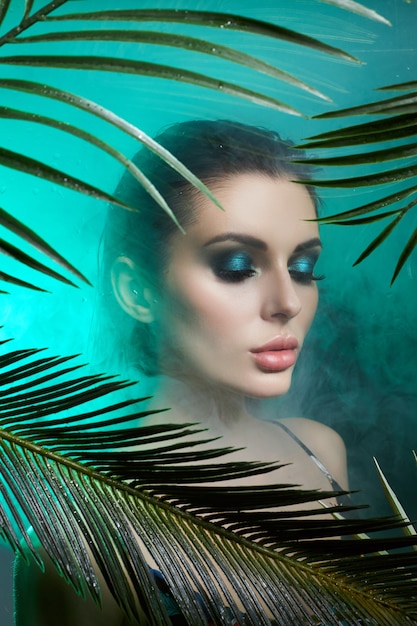 Mujer de belleza en palma deja maquillaje húmedo, retrato tropical chica en traje de baño verde en ramas de palmera en estudio, humo y gotas de lluvia sobre vidrio. Mujer sexy con maquillaje verde brillante