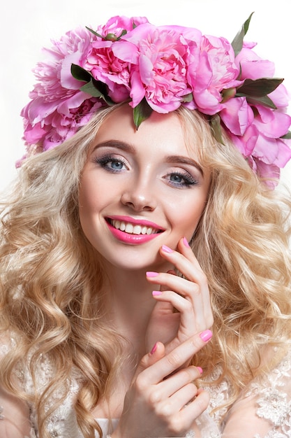 Mujer de belleza en una pared blanca. Cabello ondulado brillante y una corona de peonías rosas