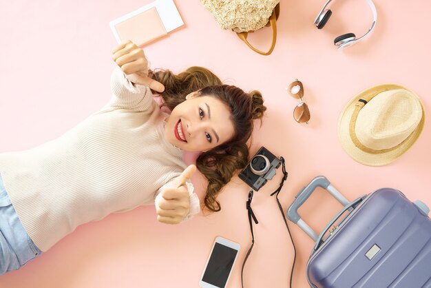 La mujer de belleza sonríe felizmente y te muestra el pulgar en el piso rosa. Acostada con su equipaje de viaje