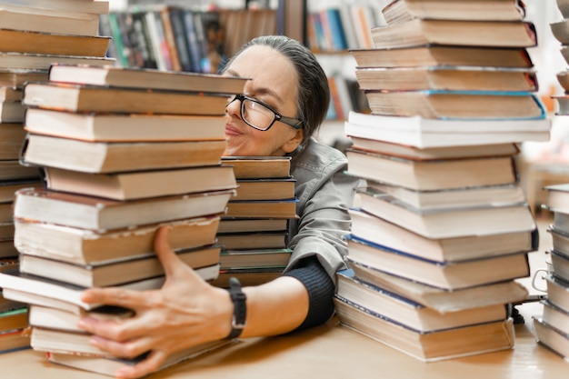 Una mujer en la biblioteca está trabajando en un escritorio. Una mujer de mediana edad con pilas de libros. Ella estudia.