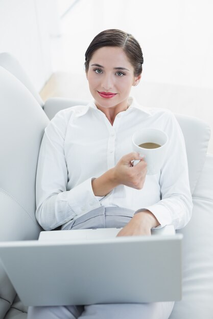 Mujer bien vestida con la taza del ordenador portátil y de café en el sofá