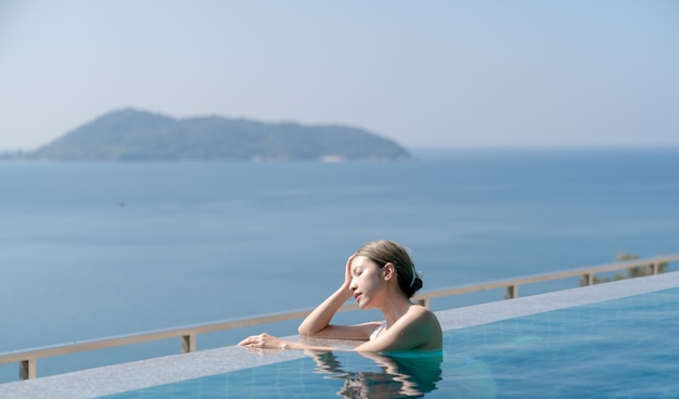 Mujer en bikini blanco relajándose en el borde de una piscina infinita con vistas al mar azul.