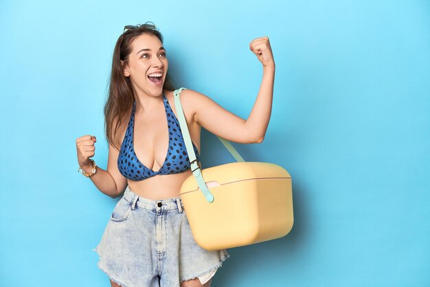 Mujer en bikini con un refrigerador de playa portátil estudio azul levantando el puño después de un concepto ganador de la victoria