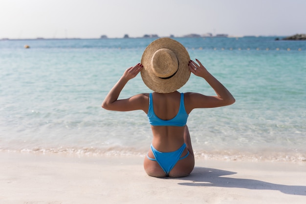 Mujer en bikini sentada y descansando en la playa con sombrero de paja