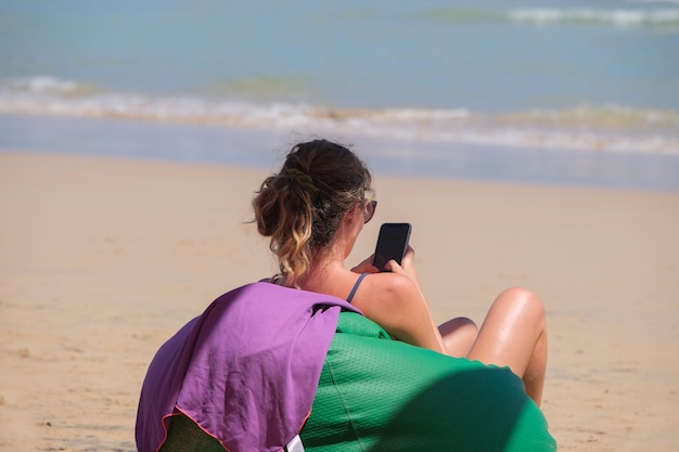 Foto mujer en bikini se sienta en una silla otomana en la playa y mira un teléfono inteligente