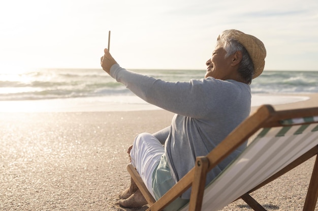 Foto mujer birracial sonriente en videollamada a través de un teléfono inteligente sentada en una silla en la playa. estilo de vida y telecomunicaciones.