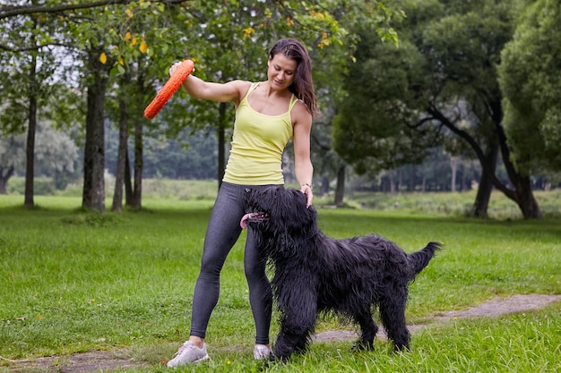 La mujer blanca está entrenando a su briard con la ayuda de un juguete mientras caminan en un parque público.