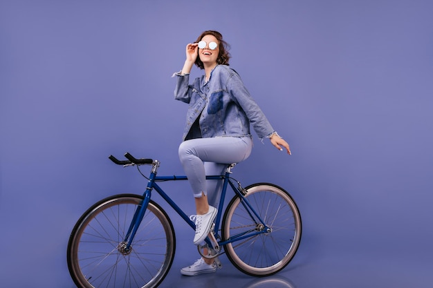 Mujer blanca inspirada sentada en bicicleta en el estudio Retrato interior de una dama segura de sí misma con cabello ondulado posando sobre fondo púrpura