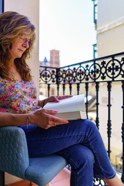 Mujer blanca madura leyendo un libro cómodamente en el balcón de su casa