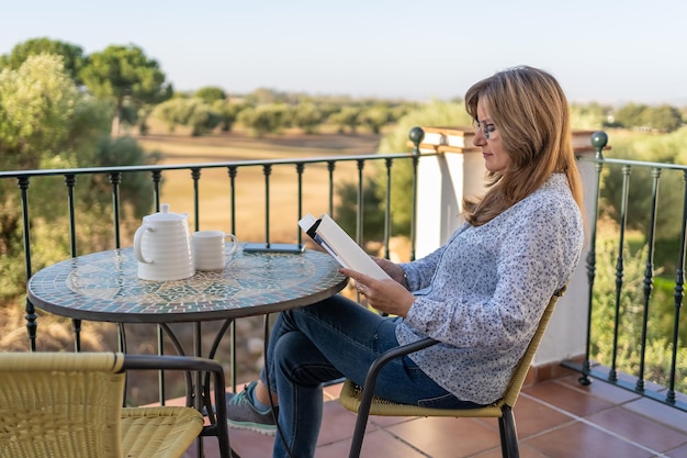 Mujer blanca madura leyendo un libro en la terraza de un apartamento con vistas al exterior en un día soleado