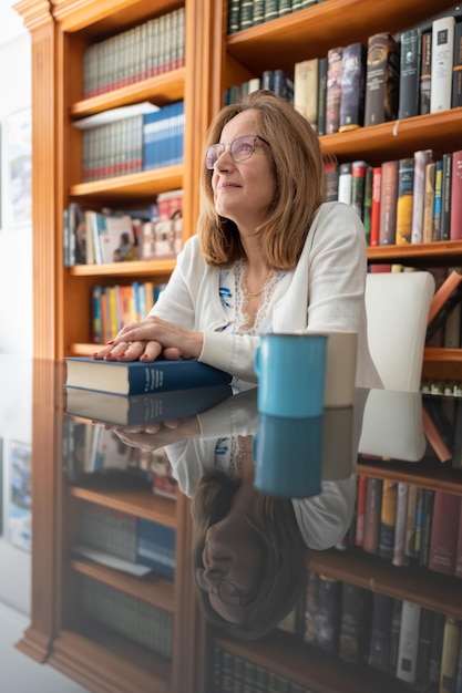 Mujer blanca mayor mirando al frente y rodeada de libros sobre una mesa de vidrio