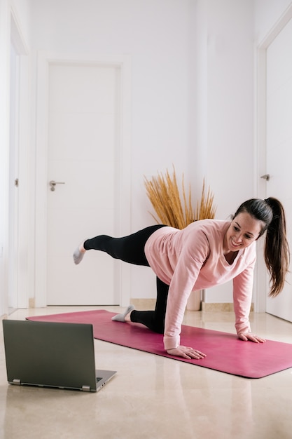 Mujer blanca de pelo oscuro entrenando en casa durante la cuarentena mientras ve la clase deportiva en la computadora portátil