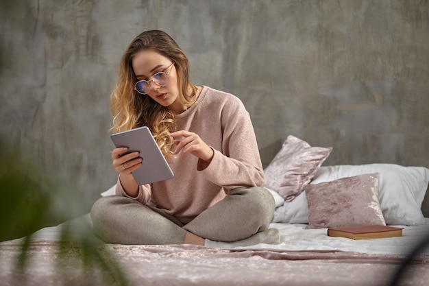 Mujer blogger en gafas ropa casual sentada en la cama en posición de loto trabajando con tableta en el dormitorio ...