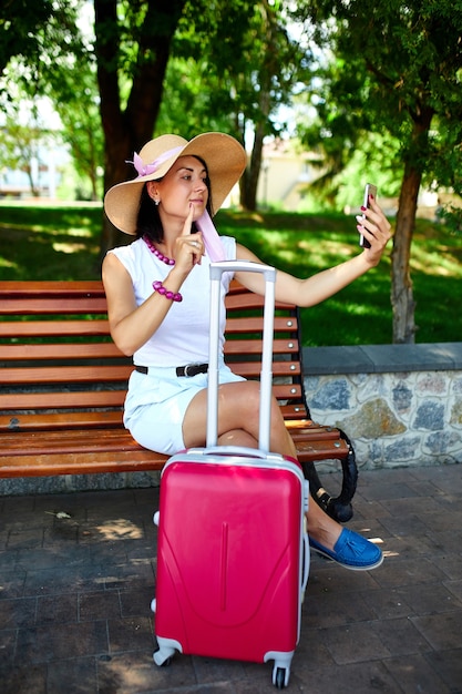 Mujer bloguera con sombrero de paja y máscara quitada se sostiene en la cabeza, en el parque al aire libre con una maleta, se toma una selfie, la vida durante la pandemia de coronavirus, la apertura de los viajes aéreos, el concepto de viaje.